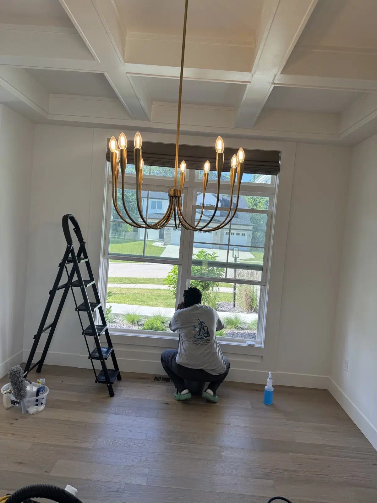 Cleaner kneeling to wash a large window beneath a brass chandelier — Leskuin Cleaning LLC in Springfield